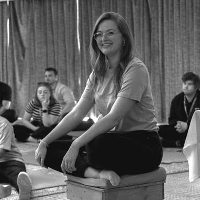 A person sitting on a stool with people in the backgroundDescription automatically generated
